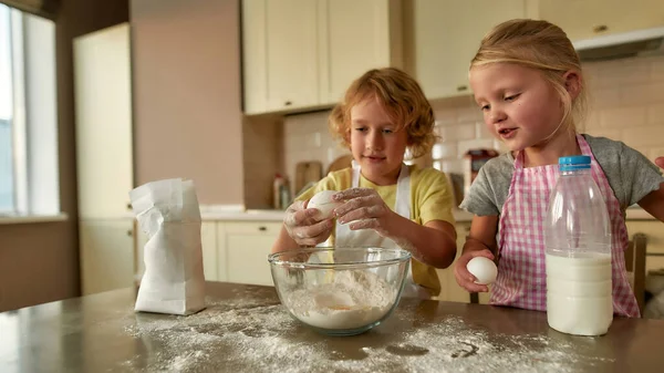 İki küçük tatlı çocuk, önlüklü kız ve erkek evde mutfak masasında hamuru birlikte hazırlarken yumurta ekliyorlar. — Stok fotoğraf