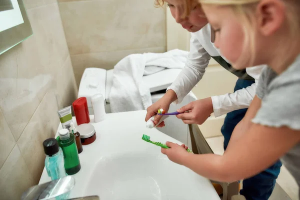 Primer plano de las manos de dos niños, niños y niñas sosteniendo cepillos de dientes, apretando la pasta de dientes del tubo mientras se cepillan los dientes en casa en el baño — Foto de Stock