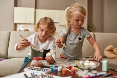 Sevimli küçük çocuklar, erkek ve kız kardeşler renkli Paskalya yumurtaları boyuyor, evdeki kanepede oturuyor.