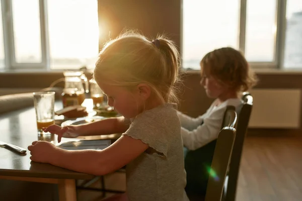 Vista lateral de la niña enfocada usando tableta PC mientras desayuna o almuerza, sentada junto a su hermano en la mesa de la cocina — Foto de Stock