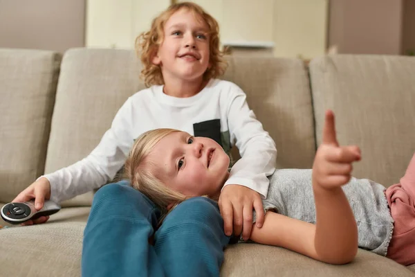 Retrato de hermanitos lindos, niño y niña mirando enfocados mientras ven la televisión juntos, abrazándose en un sofá en casa —  Fotos de Stock