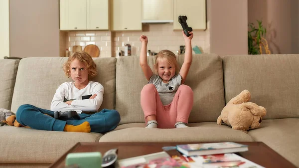 Competencia. Adorable niña que parece emocionada mientras juega videojuegos con su hermano, sentada en un sofá en la sala de estar — Foto de Stock