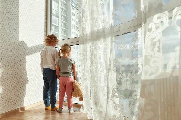 Två små barn i hemkläder som håller hand när de står inomhus och tittar ut genom ett stort fönster en solig dag — Stockfoto