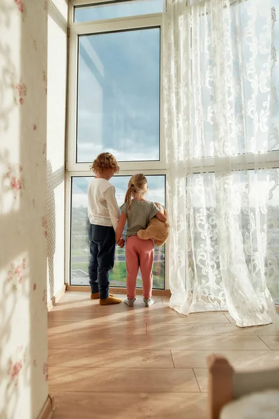 İki küçük çocuk evde el ele tutuşup pencereden kasvetli havayı izliyorlar. — Stok fotoğraf
