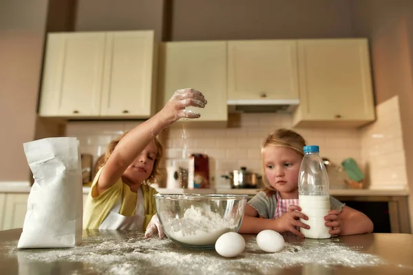 İki küçük çocuk, erkek ve kız evde mutfak masasında hamuru pişirirken un, süt ve diğer malzemeleri ekliyorlar. — Stok fotoğraf