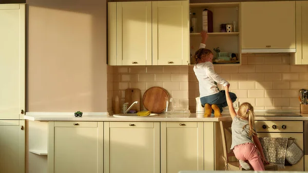 Achteraanzicht van twee stoute kinderen, broer en zus op zoek naar iets zoets in de keukenkast. klein meisje helpen haar broer door het houden van zijn hand — Stockfoto