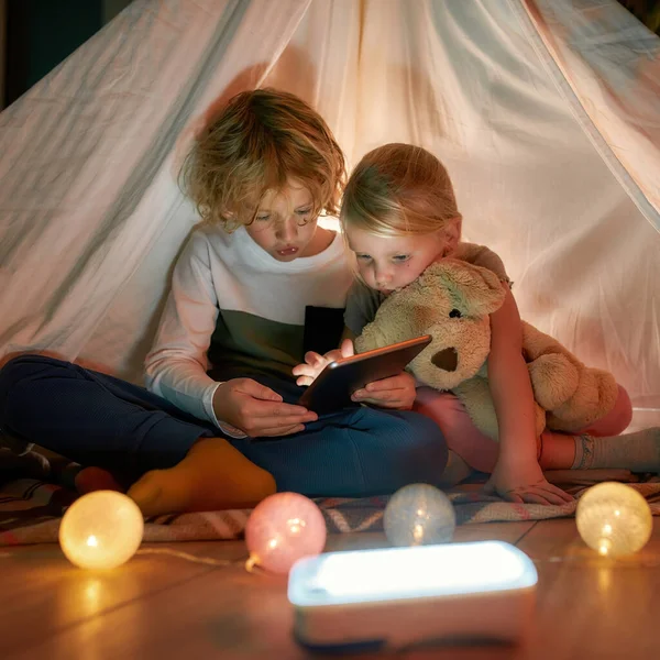 Primer plano de los niños pequeños y lindos, niño y niña usando tableta digital mientras pasan tiempo juntos, sentados en una cabaña hecha con sábanas en casa —  Fotos de Stock
