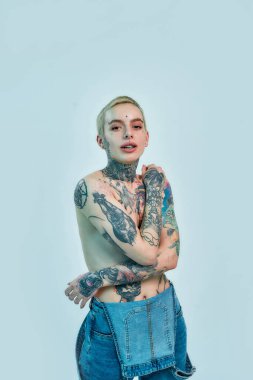 Dövme ve piercing. Beyaz üstsüz bir kadın, piercing takıyor ve dövmeleri var. Kot pantolon giymiş, elleri göğsünde, göğüslerini kapatmış kameraya bakıyor.