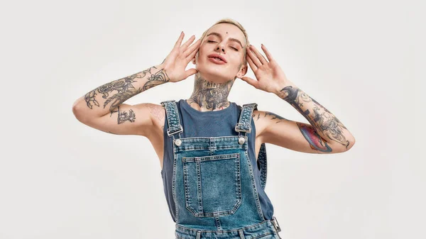 Tatoeage en piercing. Een blanke vrouw bedekt met tatoeages glimlachend met een spijkerbroek over het algemeen genieten van dansen met haar ogen dicht en handen omhoog aanraken van haar hoofd van beide kanten — Stockfoto