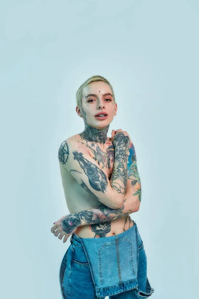 Tatuaje y piercing. Una mujer blanca en topless que tiene piercing y tatuajes mirando a una cámara mientras está de pie en un denim en general con las manos en el pecho cubriendo sus pechos — Foto de Stock