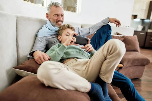 Провожу время с внуком дома. Милый мальчик играет в видеоигры на смартфоне, расслабляясь на диване со своим счастливым дедушкой — стоковое фото