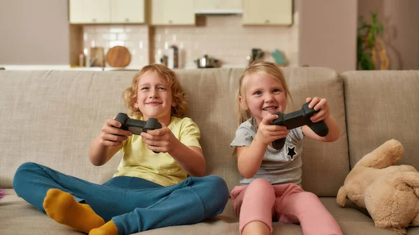 Niños emocionados, niño y niña jugando videojuegos usando joystick o controlador mientras están sentados juntos en el sofá en casa —  Fotos de Stock