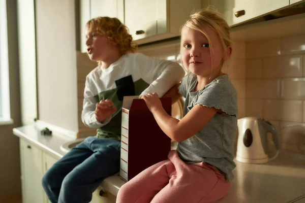 Retrato de una linda niña mirando a la cámara, comiendo copos de maíz mientras está sentada en el gabinete de la cocina junto con su hermano en casa —  Fotos de Stock