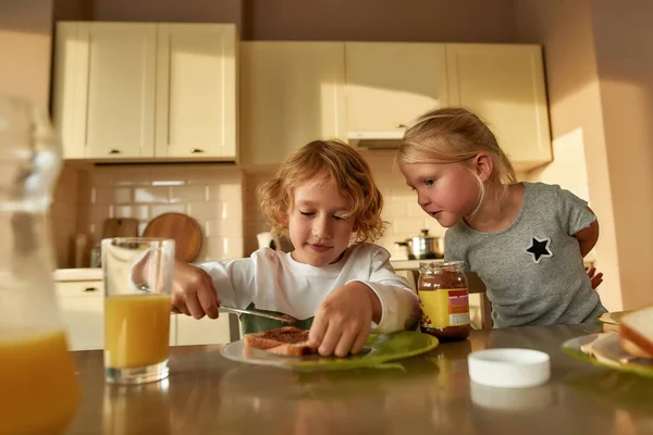 Ritratto di bambina carina che guarda suo fratello spargere burro di cioccolato sul pane tostato mentre prepara il pranzo o la colazione per sua sorella e se stesso in cucina — Foto Stock