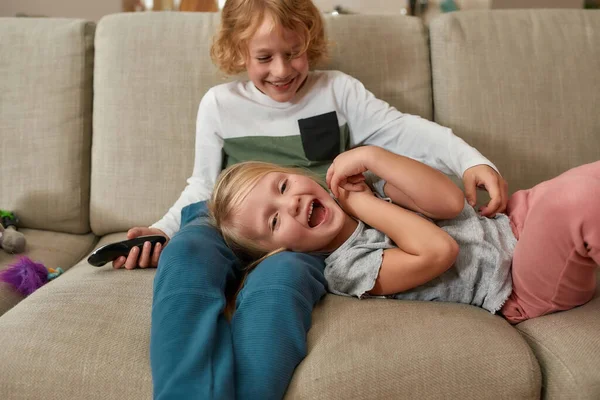 Underbara små syskon, pojke och flicka som har roligt, leker tillsammans medan de tittar på TV, gosar på en soffa hemma — Stockfoto
