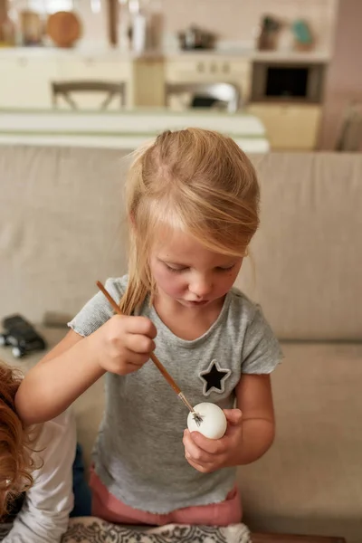 Porträtt av söt liten flicka målning färgglada påskägg, sitter på en soffa i vardagsrummet medan spendera tid hemma — Stockfoto