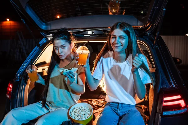 Two young women, best friends looking emotional while sitting in car trunk, having snacks and watching a movie in a drive in cinema