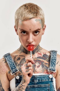 Dövme ve piercing. Kot pantolon giyen, kameraya bakarken lolipop emen beyaz, piercing yaptırmış ve dövmeli bir kadın.