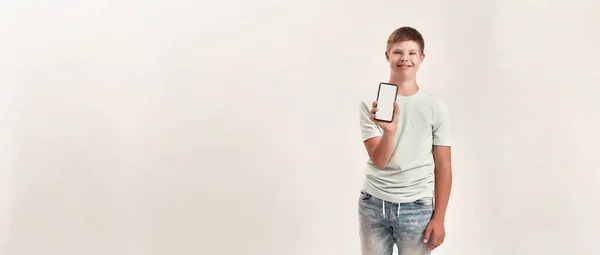 Glücklicher behinderter Junge mit Down-Syndrom lächelt, hält und zeigt Smartphone mit leerem Bildschirm vor der Kamera, während er isoliert vor weißem Hintergrund steht — Stockfoto