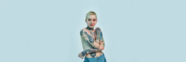 入れ墨とピアス。A white tattoed pierced woman standing topless with her hands crossed on her chest covering her breasts — ストック写真