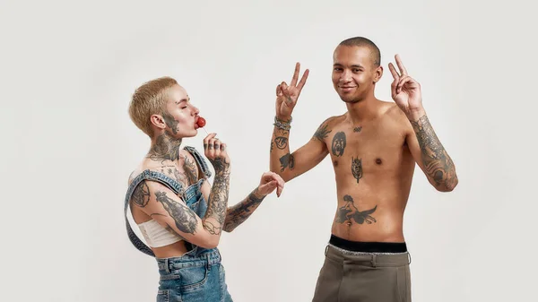 Multikulturellt förhållande. En mörkhyad topless med tatueringar och en vit tatuerad piercad tjej som båda dansar och roar sig medan hon suger en klubba. — Stockfoto