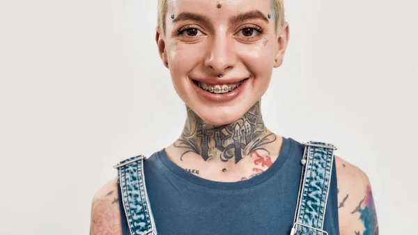 Συναισθήματα και στάσεις. Κοντινό πλάνο μιας λευκής γυναίκας με τατουάζ με σκουλαρίκια κοιτάζοντας μέσα σε μια κάμερα χαμογελώντας φορώντας παρένθεση — Φωτογραφία Αρχείου