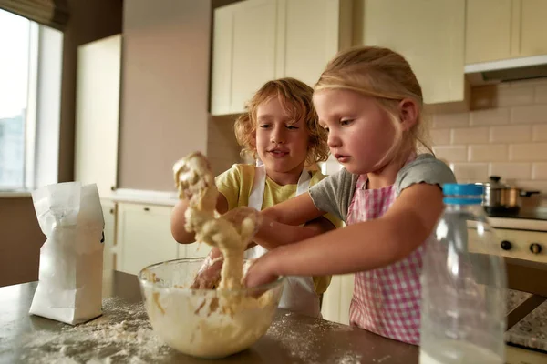 Sevimli küçük çocuklar, önlüklü kız ve erkek masada hamuru hazırlarken mutlu görünüyorlar. Çocuklar mutfakta kurabiye yaparken eğleniyor. — Stok fotoğraf