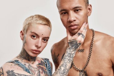 Çok kültürlü bir ilişki. Siyah derili, üstsüz bir erkeğe dokunan dövmeli, piercingli, beyaz bir kadının kameraya bakarken yakın çekimi.