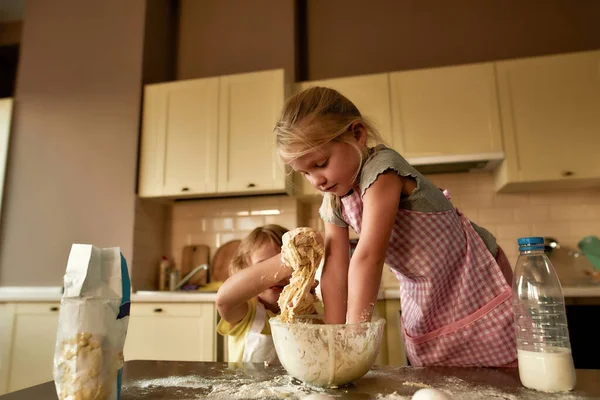 Evdeki mutfak masasında kardeşiyle kurabiye yaparken önlüklü tatlı bir kız hamur yoğuruyor. — Stok fotoğraf