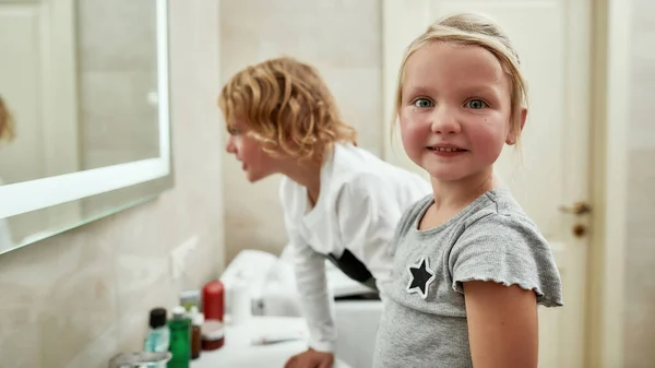 화장실에서 동생과 함께 양치질을 하면서 카메라 앞에서 웃고 있는 귀여운 어린 소녀의 모습 — 스톡 사진