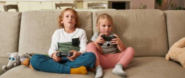 İki kardeş, küçük bir oğlan ve kız odaklanmışlar, evde bir kanepede otururken oyun oynuyorlar.