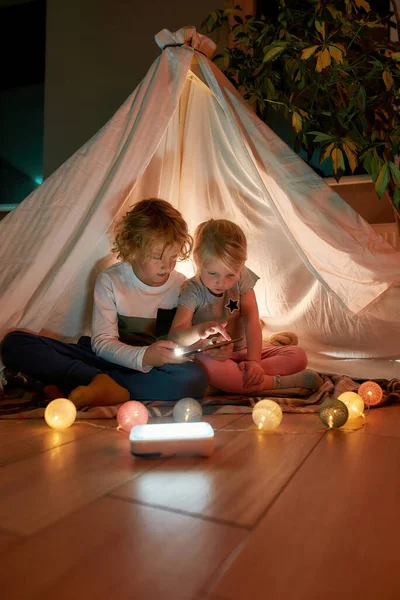Dos niños pequeños, hermano y hermana usando tableta PC mientras están sentados en una manta en un tipi hecho con sábanas en casa —  Fotos de Stock