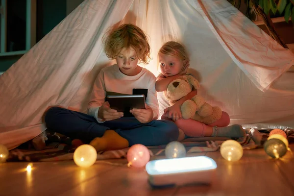 Adorable niño usando tableta PC mientras pasa tiempo con su hermana pequeña, sentado en una manta en una cabaña hecha con sábanas en casa —  Fotos de Stock