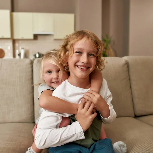 Найкращі друзі. Портрет щасливих братів і сестер, маленький хлопчик і дівчинка посміхаються на камеру, обіймаючи один одного, сидячи на дивані вдома — стокове фото