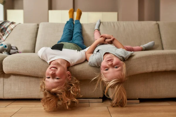 Porträtt av lekfulla barn, liten pojke och flicka leende medan liggande upp och ner på en soffa i vardagsrummet — Stockfoto