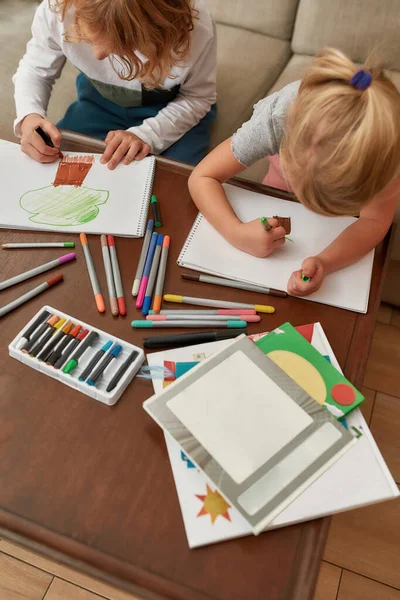 Hög vinkel syn på fokuserade små barn, pojke och flicka ritning på papper med markör penna, sitter tillsammans på en soffa hemma — Stockfoto