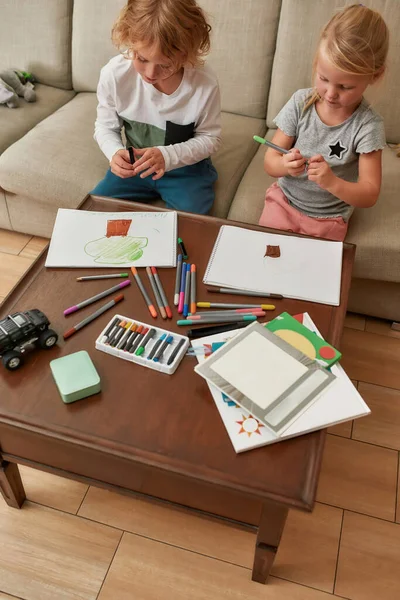 Високий кут зору чарівних маленьких дітей, хлопчик і дівчинка малюють на папері за допомогою маркерної ручки, сидячи разом на дивані вдома — стокове фото