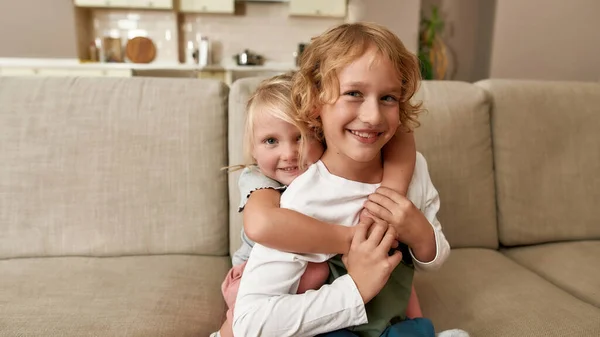 Портрет радісних братів і сестер, маленький хлопчик і дівчинка посміхаються на камеру, обіймаючи один одного, сидячи на дивані вдома — стокове фото