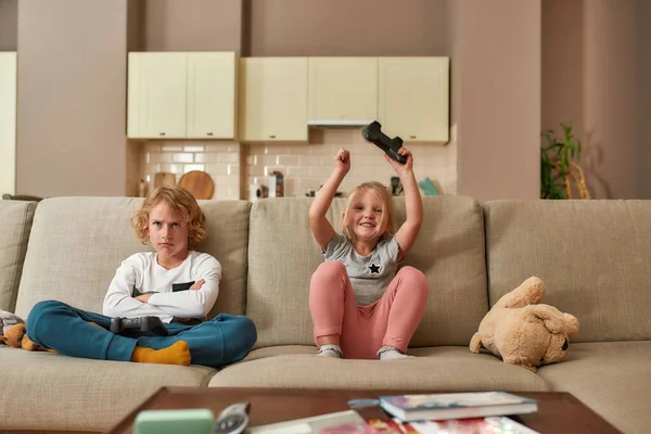 Eu ganhei. Adorável menina olhando animado enquanto joga jogos de vídeo com seu irmão, sentado em um sofá na sala de estar — Fotografia de Stock