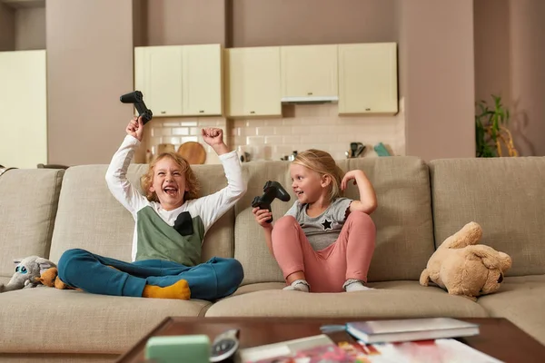 Niños felices. Adorables niños pequeños mirando emocionados mientras juegan videojuegos juntos, sentados en un sofá en la sala de estar —  Fotos de Stock