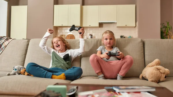 Dos hermanos lindos, niño y niña jugando videojuegos, sentados en un sofá en la sala de estar —  Fotos de Stock