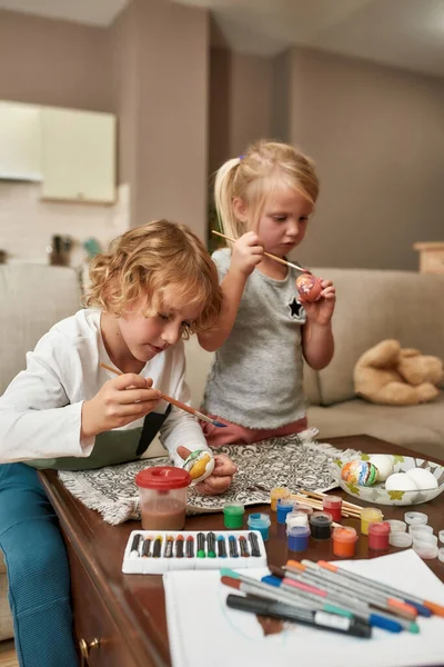 Retrato de dos niños pequeños, hermano y hermana mirando enfocados mientras pintan coloridos huevos de Pascua, sentados en un sofá en casa — Foto de Stock