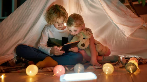 Adorables niños pequeños, niños y niñas usando tabletas digitales mientras pasan tiempo juntos, sentados en una cabaña hecha con sábanas en casa —  Fotos de Stock