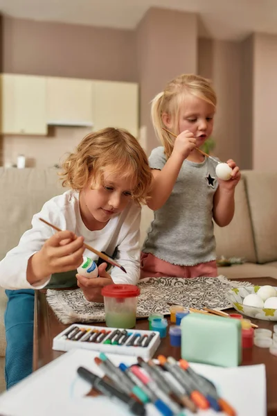 Roztomilé malé děti, chlapec a dívka malování barevné velikonoční vajíčka v obývacím pokoji, zatímco tráví čas spolu doma — Stock fotografie