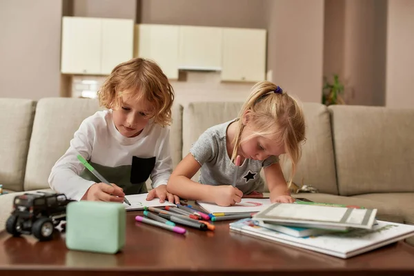 Crea. Lindos niños pequeños, niño y niña mirando enfocados mientras dibujan en papel usando rotulador, sentados juntos en un sofá en casa — Foto de Stock