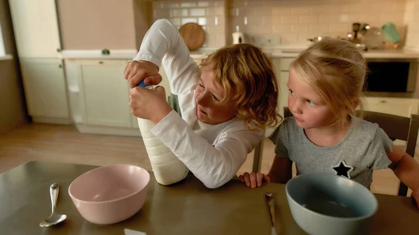 Tatlı çocuk kaseye koymak için bir şişe süt açmaya çalışıyor. İki küçük çocuk kahvaltı için hazırlanıyor, mutfaktaki masada birlikte oturuyorlar. — Stok fotoğraf