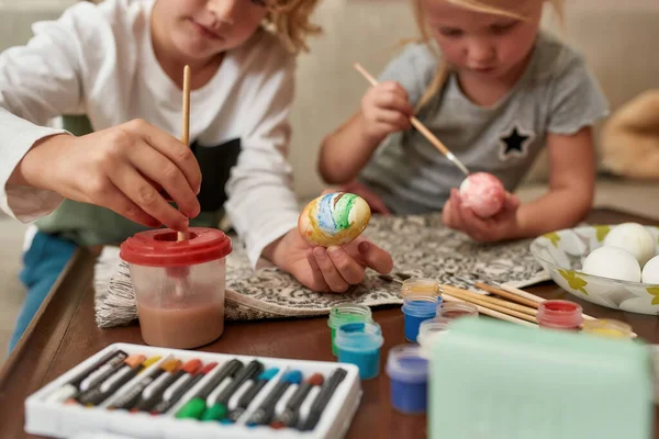 Detailní záběr na ruce rozkošný chlapeček malování barevné velikonoční vajíčka spolu s jeho sestrou, zatímco tráví čas doma — Stock fotografie