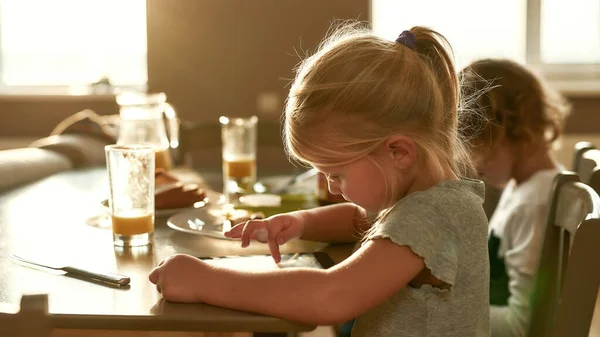 Retrato de chica bonita enfocada usando tableta PC mientras desayunaba o almorzaba, sentada junto a su hermano en la mesa de la cocina —  Fotos de Stock