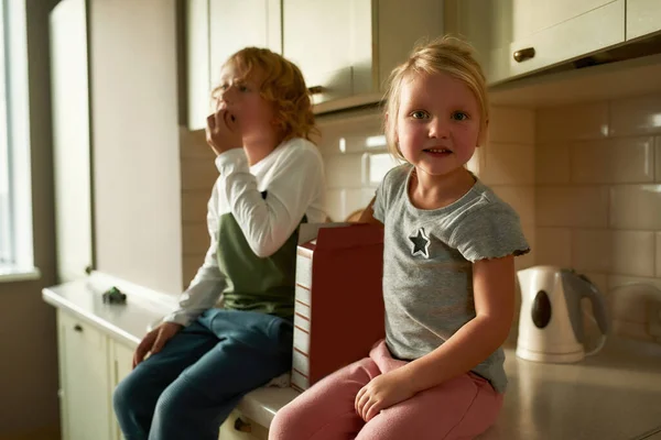 カメラで笑っている可愛い女の子の肖像画,家で台所のキャビネットに座っている間、彼女の兄と一緒にコーンフレークを食べます — ストック写真