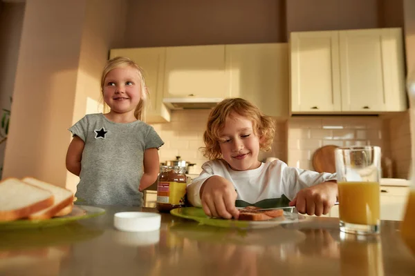 Küçük şirin çocukların portresi kendilerine öğle yemeği ya da kahvaltı hazırlıyorlar. Neşeli çocuk kızarmış ekmeğe çikolata fındık ezmesi sürüyor. — Stok fotoğraf